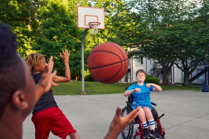 Three teenage friends play basketball at a neighborhood park. One of the teens, a Black boy with short natural black hair styled high on top, passes the ball to another teen, a white boy seated in a manual wheelchair. A third teen, a light-skinned African American male, runs towards the hoop. The teen seated in the wheelchair has a gentle smile on his face and is wearing blue athletic wear. The African American male has is wearing black and red athletic wear and a gold chain necklace.