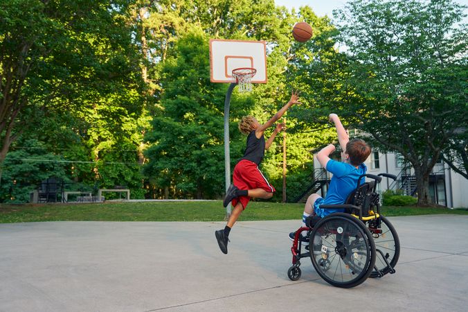 Two teens play basketball at a neighborhood park. One teen, a white boy seated in a manual wheelchair, shoots the ball. The other teen, a light-skinned African American male, jumps forward with arms outstretched, defending the shot. The teen seated in a manual wheelchair has short brown hair and is wearing blue athletic wear. The other teen is wearing black and red athletic wear.