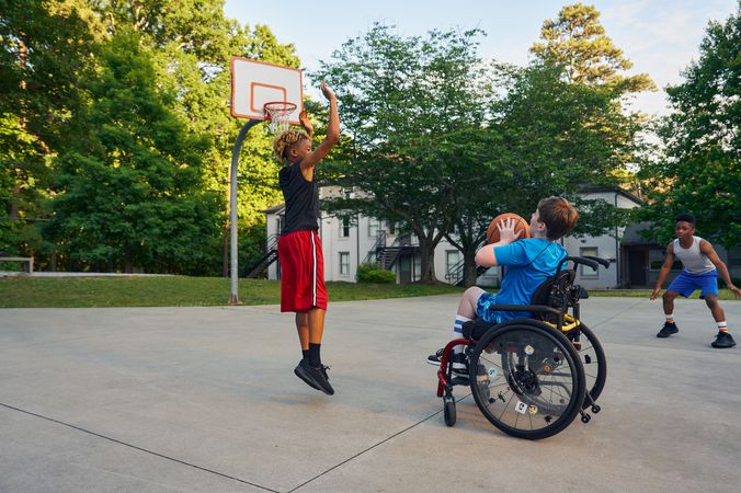Three teens play basketball at a neighborhood park. One teen, a white boy seated in a manual wheelchair, holds the ball, ready to shoot. A second teen, a light-skinned African American male, jumps forward with arms outstretched, ready to defend the shot. A third teen, a Black boy, crouches, ready to receive a pass. The teen seated in a manual wheelchair has short brown hair and is wearing blue athletic wear. The second teen defending has curly brown to blonde hair is wearing black and red athletic wear. The third teen has short natural black hair styled high on top and is wearing gray and blue athletic wear.
