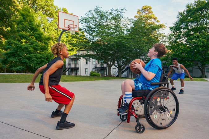 Three teens play basketball at a neighborhood park. One teen, a white boy seated in a manual wheelchair, holds the ball, ready to shoot. A second teen, a light-skinned African American male, crouches in a jumping position. A third teen, a Black boy, crouches, ready to receive a pass. The teen seated in a manual wheelchair has short brown hair and is wearing blue athletic wear. The second teen ready to jump has curly brown to blonde hair is wearing black and red athletic wear. The third teen has short natural black hair styled high on top and is wearing gray and blue athletic wear.