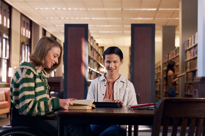 A white teenage girl seated in a manual wheelchair smiles while reading a book at a table in a library. Next to her, a Chinese teen, who is a blind cane user, reads on a refreshable braille display. His white cane is collapsed and is placed the table next to him. The teen girl seated in a manual wheelchair has a dark blonde bob and is wearing a green and white striped knit shirt and jeans. The teen boy has slicked back black hair and a friendly smile with braces. He is wearing a rust colored shirt under a white plaid button down shirt.