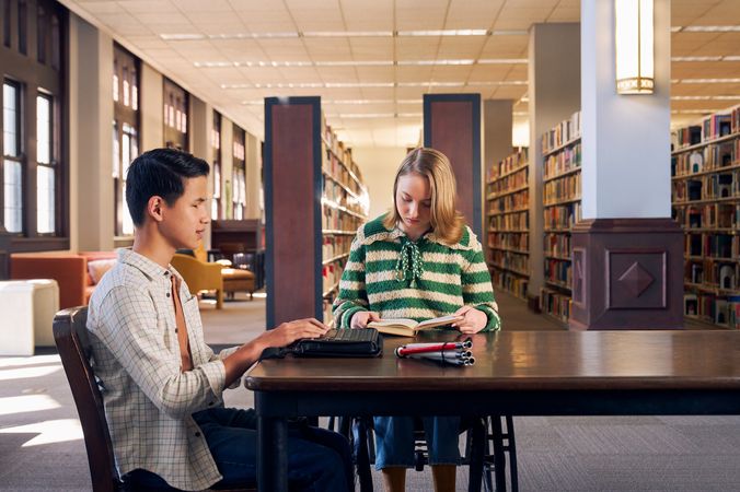 Two teenage friends read at a table in a library. One teen, a Chinese teen, who is a blind cane user, reads on a refreshable braille display. His white cane is collapsed and is placed the table next to him. The other teen, a white teenage girl seated in a manual wheelchair, is seated in a manual wheelchair and holds a hardcover book open. The teen girl has a dark blonde bob and is wearing a green and white striped knit shirt and jeans. The teen boy has slicked back black hair and is wearing a white plaid button down shirt.