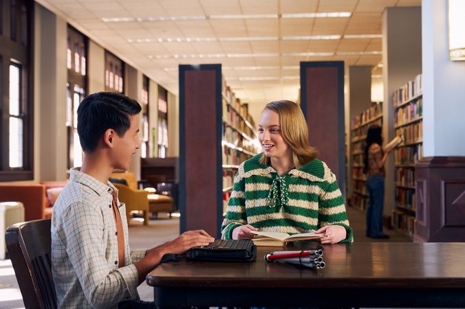 Two teenage friends smile and chat at a table in a library. One teen, a Chinese teen, who is a blind cane user, has a refreshable braille display in front of him. His white cane is collapsed and is placed the table next to him. The other teen, a white teenage girl seated in a manual wheelchair, is seated in a manual wheelchair and holds a hardcover book open. The teen girl has a dark blonde bob and is wearing a green and white striped knit shirt and jeans. The teen boy has slicked back black hair and is wearing a white plaid button down shirt.
