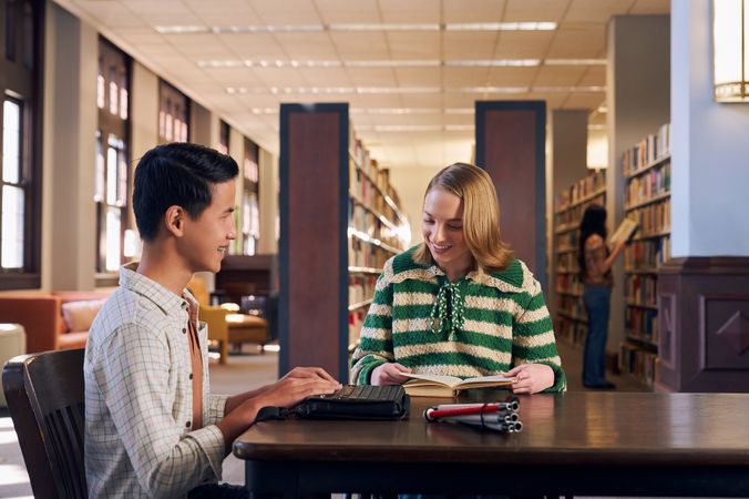 Two teenage friends laugh and read at a table in a library. One teen, a Chinese teen, who is a blind cane user, reads on a refreshable braille display. His white cane is collapsed and is placed the table next to him. The other teen, a white teenage girl seated in a manual wheelchair, is seated in a manual wheelchair and looks down at a hardcover book she has open. The teen girl has a dark blonde bob and is wearing a green and white striped knit shirt and jeans. The teen boy has slicked back black hair and is wearing a white plaid button down shirt.