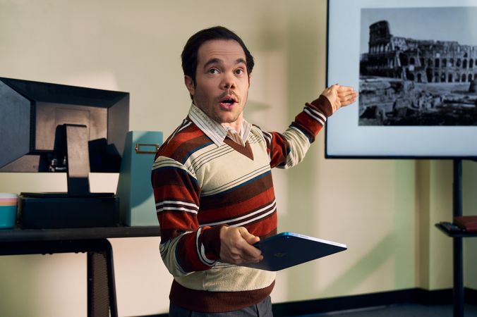 A white male professor with medium brown hair and light brown eyes who has dwarfism gestures at a large monitor next to him, displaying a black and white photo of the Colosseum. He is holding a tablet in his other hand. He is dressed in a striped V-neck sweater, charcoal dress pants, and dressy-casual shoes, standing with a warm, confident presence. Behind him, his desk has a computer, a file holder, and a mug.