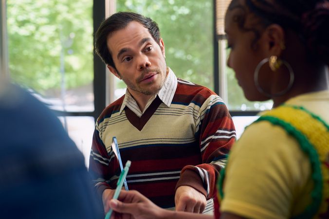 A white male professor with medium brown hair and light brown eyes who has dwarfism stands and talks to a student with focus. He is holding a tablet with one hand and taps the student's desk with the other. The student takes notes as the professor speaks. The professor is wearing a striped V-neck sweater, charcoal dress pants, and dressy-casual shoes. The student is a Black woman with hair pulled into a bun and is wearing a yellow shirt. Behind them are large windows that show a college campus.