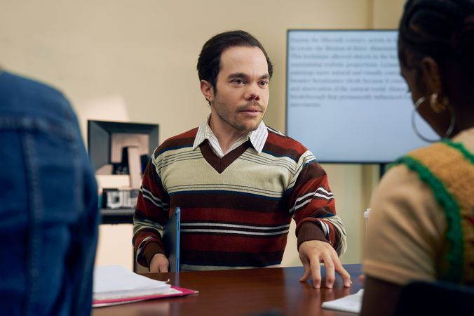 A white male professor with medium brown hair and light brown eyes who has dwarfism stands and talks to a student with focus. He is holding a tablet with one hand and taps the student's desk with the other. The student takes notes as the professor speaks. The professor is wearing a striped V-neck sweater, charcoal dress pants, and dressy-casual shoes. The student is a Black woman with hair pulled into a bun and is wearing a yellow shirt. Behind them is a large monitor with text displayed.