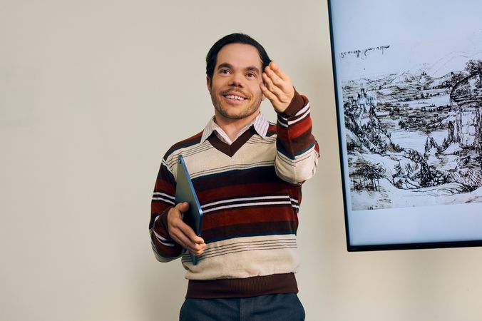 A white male professor with medium brown hair and light brown eyes who has dwarfism smiles and gestures to a student off camera, calling on them. Next to him is a large monitor displaying a pen and ink drawing by Leonardo da Vinci. He is holding a tablet in his other hand. He has short brown hair and is wearing a red striped sweater, a collared shirt, and dark dress pants.