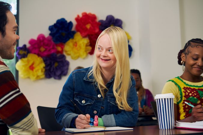 A caucasian college student with Down Syndrome sits at a lecture table, smiles, and chats with her professor, who is standing next to her. She has long blonde hair, blue eyes, and is wearing a jean hacket and several bright scrunchies on her wrist. Another student next to her, a Black woman with braided hair pulled into a bun, writes in a notebook. A large flower sculpture hangs on the wall behind her. Her professor, a white man with medium brown hair and light brown eyes who has dwarfism, smiles and leans on the students' lecture table.