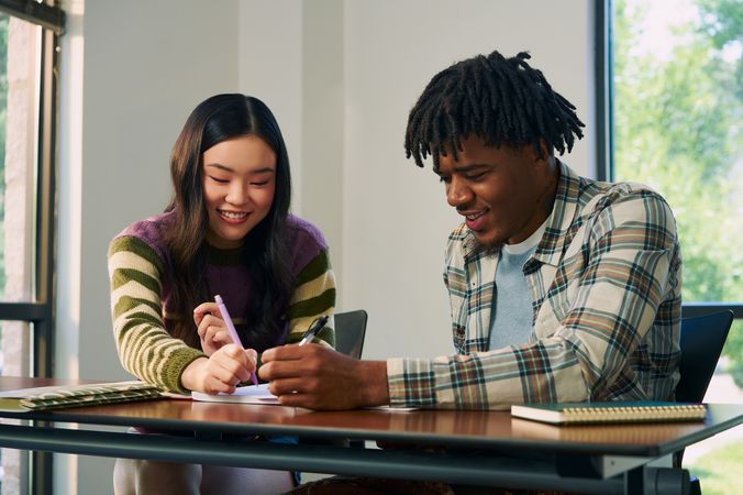 Two college students smile and write in a worksheet together at a lecture table. One student, a Black man, has short locs and is wearing a white plaid shirt and a blue knit shirt underneath. The other student, an East Asian young woman, has long curled hair and is wearing a purple, green, and tan striped sweater.