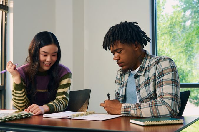 Two college students study notes in an open notebook together at a lecture table. They are both holding pencils. One student, a Black man with short locs is wearing a white plaid shirt and a blue knit shirt underneath. The other student, an East Asian young woman, has long curled hair and is wearing a purple, green, and tan striped sweater.