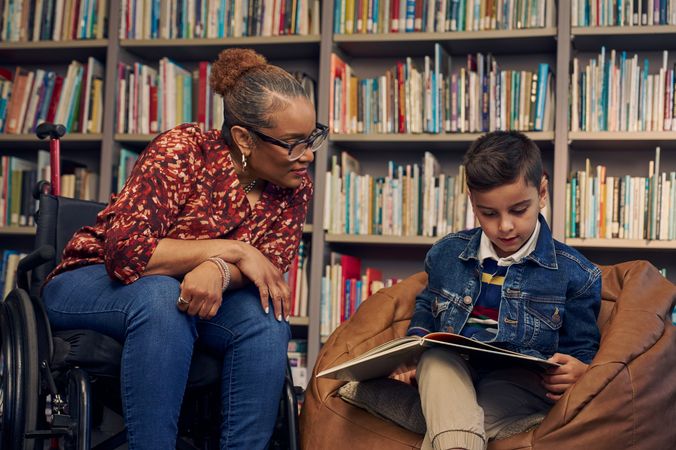 An African American teacher seated in a manual wheelchair watches a young student sitting in a bean bag chair read a picture book. The teacher has medium brown skin, reddish brown hair pulled into a short natural ponytail, and is wearing reading glasses, a red patterned blouse, and jeans. The young student is a Hispanic and Mediterranean-American boy with short brown hair. He has olive skin and is wearing a blue striped shirt, a jean jacket, and khaki pants. A wall of children's books are behind them.