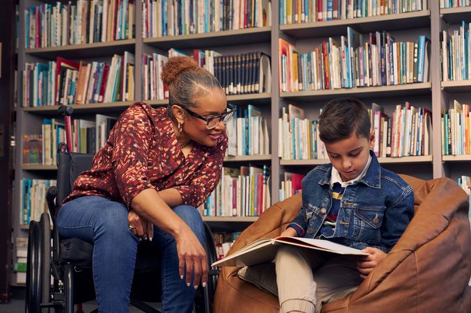 An African American teacher seated in a manual wheelchair smiles and watches a young student sitting in a bean bag chair read a picture book. The teacher has medium brown skin, reddish brown hair pulled into a short natural ponytail, and is wearing reading glasses, a red patterned blouse, and jeans. The young student is a Hispanic and Mediterranean-American boy with short brown hair. He  has olive skin and is wearing a blue striped shirt, a jean jacket, and khaki pants. A wall of children's books are behind them.