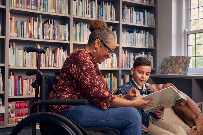 A young boy sitting in a bean bag chair and his African American teacher seated in a manual wheelchair hold and look at a picture book together. The teacher has a red walking cane fastened to the back of her wheelchair. She has medium brown skin, reddish brown hair pulled into a short natural ponytail, and is wearing reading glasses, a red patterned blouse, and jeans. The young student is a Hispanic and Mediterranean-American boy with short brown hair. He  has olive skin and is wearing a blue striped shirt, a jean jacket, and khaki pants. A wall of children's books are behind them.