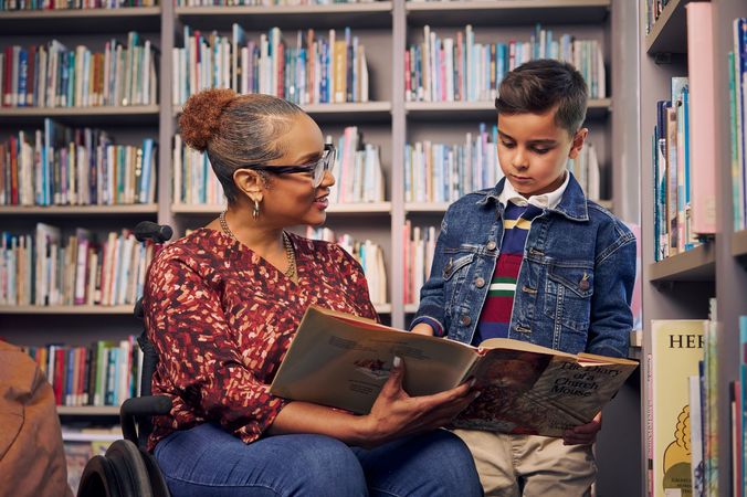 An African American teacher seated in a manual wheelchair holds a picture book open while her young student reads out loud. The teacher has medium brown skin, reddish brown hair pulled into a short natural ponytail, and is wearing reading glasses, a red patterned blouse, and jeans. The young student is a Hispanic and Mediterranean-American boy with short brown hair. He  has olive skin and is wearing a blue striped shirt, a jean jacket, and khaki pants. A wall of children's books are behind them.