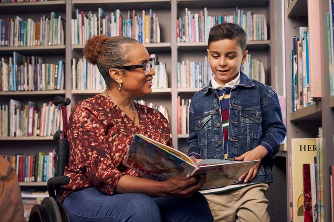 An African American teacher seated in a manual wheelchair smiles and holds a picture book open while her young student reads out loud. The teacher has medium brown skin, reddish brown hair pulled into a short natural ponytail, and is wearing reading glasses, a red patterned blouse, and jeans. The young student is a Hispanic and Mediterranean-American boy with short brown hair. He  has olive skin and is wearing a blue striped shirt, a jean jacket, and khaki pants. A wall of children's books are behind them.